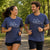 A man and a woman jogging side-by-side on a park path, wearing matching heather navy blue t-shirts with a white sneaker graphic and the text '26.2' printed on the chest.