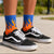 A person wearing black Vans sneakers and bright blue crew socks with a yellow and orange flame pattern, standing outdoors on a paved surface with a blurred green background.