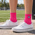Bright pink crew socks with embroidered soccer ball worn with white Vans sneakers on a concrete surface at an outdoor soccer field.