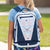 A blonde girl seen from behind wearing a light blue t-shirt and a colorful patterned skirt, carrying a white and navy blue hockey-themed backpack outdoors on a sunny day.