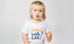 Child wearing a white lacrosse baby onesie with "Crawl Walk Lax" text and a small lacrosse stick graphic.