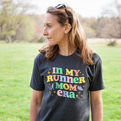 Woman wearing gray running t-shirt featuring colorful "In My Runner Mom Era" text with retro design.