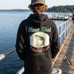 Person wearing a black hoodie with 'Hook Shot Fishing Club' design by a lake.