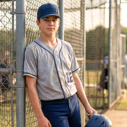 Young baseball player in uniform leaning against a fence on a field