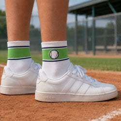 White sneakers worn with white crew socks featuring a green band, navy stripes, and a baseball graphic, photographed on a baseball field.