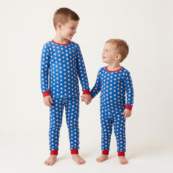 Two young boys standing and holding hands, wearing matching long-sleeve pajamas with a blue base, white baseball pattern, and red trim at the collar, cuffs, and ankles, against a plain light background.