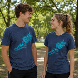 A man and a woman standing outdoors on a park path, both wearing heathered navy blue t-shirts with a bright teal graphic of a winged foot filled with inspirational running text.