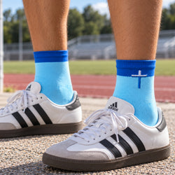 A close-up of a person's legs wearing light blue crew socks with a white cross on the cuff, paired with white Adidas sneakers on an outdoor track field.