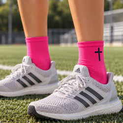 Bright pink crew socks worn with white Adidas running shoes on a green turf field. One sock features a navy cross graphic near the cuff.