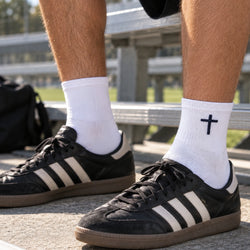 A close-up of a person's legs wearing black Adidas sneakers with white socks featuring a navy cross on the ankle, set against a blurred outdoor stadium background.