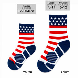 A pair of crew socks with an American flag pattern, featuring red and white stripes, a navy blue cuff with white stars, and navy blue heels and toes, displayed on a solid white background.