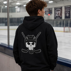 A man viewed from behind wearing a black hoodie with a white graphic of a hockey player skull on the back, standing at the boards of an ice rink.