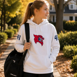 A young woman wearing a white pullover hoodie with a red wildcat mascot logo on the chest and navy blue numbers on the sleeves, carrying a black Bauer duffel bag on a sunny outdoor path.