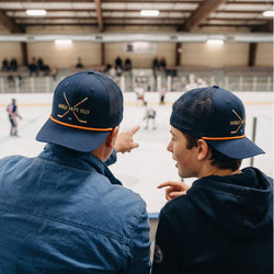 Two people wearing blue caps with dangle, snip, celly text sitting on bleachers at an indoor sports arena.