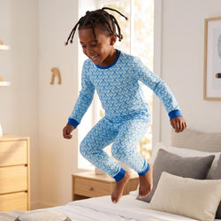 A young child wearing a light blue and white hockey patterned long-sleeve pajama set jumps onto a bed in a bright bedroom.