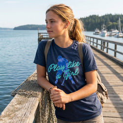Girl wearing a blue t-shirt with a hockey girl graphic and text, standing on a wooden dock by a body of water.