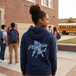 Girl wearing a hoodie with 'Play like a girl' text and graphic, standing in front of a school building with a yellow bus in the background.