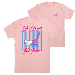 Pink t-shirt with hockey graphic and text 'She Shoots She Scores' on a white background
