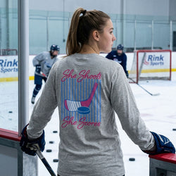 Person wearing a gray long-sleeve shirt with 'She Shoots, She Scores' design on an ice rink.