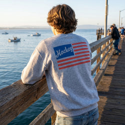 Person wearing a gray sweatshirt with 'Hockey' and an American flag design, standing on a wooden pier by the water.