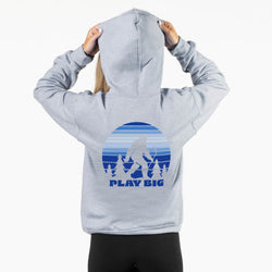 A person shown from the back wearing a light heather gray hoodie with a blue graphic print on the back. The graphic features a Bigfoot silhouette inside a striped sunset circle above a mountain range and the text 'PLAY BIG'.