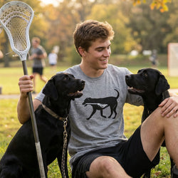 Boy sitting with two black dogs holding a lacrosse stick in a park setting