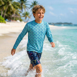 Child in a blue lacrosse patterned rash guard and dark shorts playing in ocean waves on a beach.