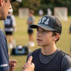Two individuals on a sports field, one wearing a cap with a lacrosse design, in an outdoor setting.