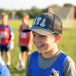 Young boy wearing a lacrosse cap and sports jersey on a field with other children in the background.