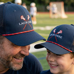 Man and young boy wearing lacrosse-themed hats on a sports field.