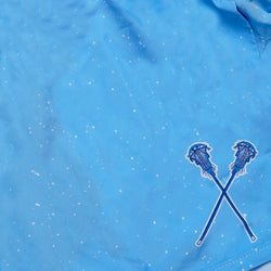 A close-up of a light blue fabric surface with small white speckles and a small embroidered graphic in the lower right showing two crossed blue-lacrosse sticks outlined in white.