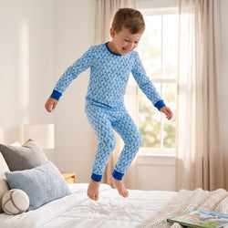 A young boy wearing a light blue and white lacrosse patterned long-sleeve pajama set jumps on a white bed in a bright bedroom.