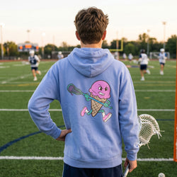 Person wearing a light blue hoodie with a pink ice cream cone holding a lacrosse stick on a sports field.