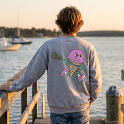 Person wearing a sweatshirt with a colorful ice cream lacrosse graphic design by a waterfront at sunset.