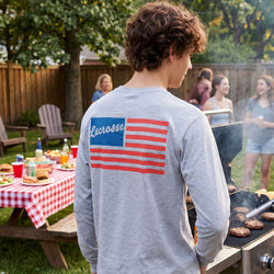 Person wearing a gray shirt with an American flag design, standing at a grill in a backyard setting.