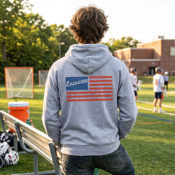 Person wearing a gray hoodie with 'Lacrosse' and an American flag design on a sports field.
