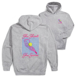 Two heather gray hoodies shown on a white background, with the back view in front and the front view behind. The back hoodie features a large graphic with pink text reading 'She Shoots She Scores,' a pink lacrosse stick, a yellow ball, and vertical blue stripes.