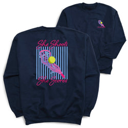 A navy blue crewneck sweatshirt shown from both the front and back. The back features a large graphic with pink text reading 'She Shoots She Scores' and a stylized lacrosse stick holding a yellow ball in front of vertical blue stripes.