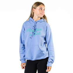 A young woman modeling a light blue hoodie with a graphic on the front that reads 'Play like a Girl' in teal script with a blue gymnast illustration. She is wearing black leggings against a plain white studio background.