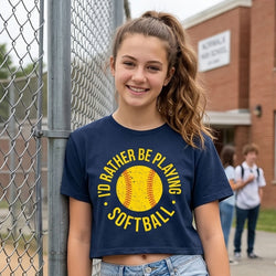 Girl wearing a navy blue t-shirt with a softball graphic and text, standing near a chain-link fence.