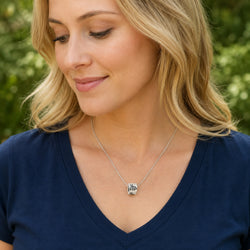 A blonde woman wearing a silver pendant necklace with a black and white zebra design, paired with a navy blue V-neck top, set against a soft-focus green outdoor background.