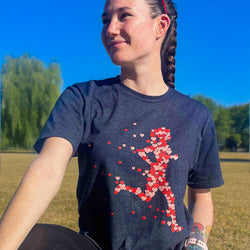 A woman wearing a dark heather gray t-shirt with a red and pink heart-patterned running figure graphic, photographed outdoors in a grassy park under a blue sky.