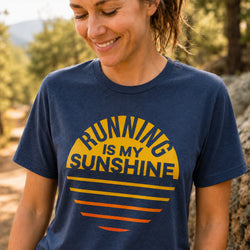 A woman outdoors wearing a navy blue graphic t-shirt with a yellow and orange sunset design and the text 'RUNNING IS MY SUNSHINE'.