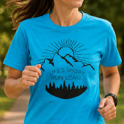 A woman wearing a bright blue short-sleeve t-shirt with a black graphic of mountains, a sun, trees, and the text 'LIFE'S SHORT RUN LONG', shown in an outdoor park setting.