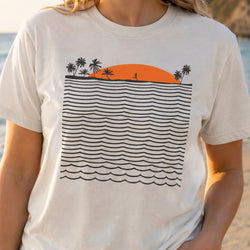 A person wearing a white short-sleeve T-shirt with a graphic print of a black wavy ocean pattern, a black palm tree silhouette, and an orange semi-circle sunset.