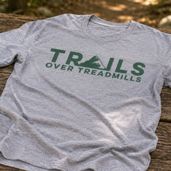 A heather gray short-sleeve t-shirt with a green graphic reading 'TRAILS OVER TREADMILLS' and a mountain icon, laid flat on a rustic wooden surface with a blurred outdoor background.