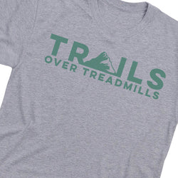 A close-up of a heather gray t-shirt featuring a green graphic print that reads 'TRAILS OVER TREADMILLS' with a mountain silhouette integrated into the lettering.