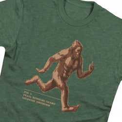 A close-up of a heather green t-shirt featuring a graphic print of Bigfoot running, with the text 'Trail Running Champ Location Unknown'