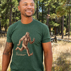 A man wearing a dark green heather graphic t-shirt with a brown Bigfoot print, standing outdoors in a forest setting.