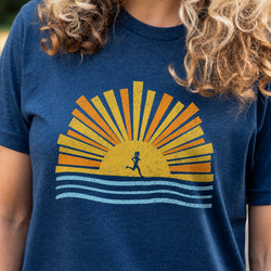 A close-up of a person wearing a heather navy blue t-shirt with a retro-style graphic of a running silhouette at sunset, featuring yellow, orange, and light blue rays.